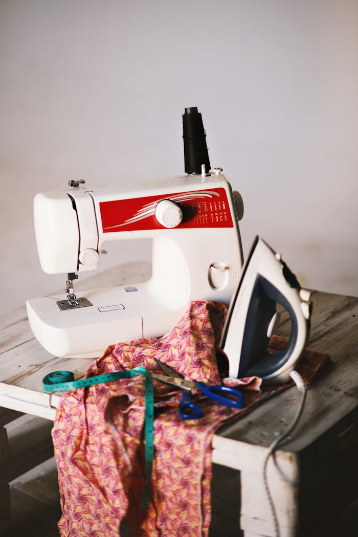 Close-up of a sewing setup with machine, iron, fabric, and tools on a wooden table.
